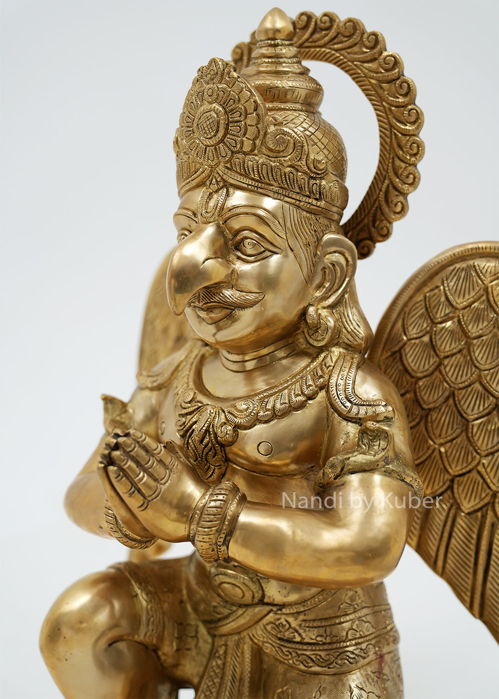 Handcrafted brass Garuda sitting statue with folded hands, detailed wings, ornate crown, and round pedestal, ideal for home temples, spiritual décor, and sacred spaces.