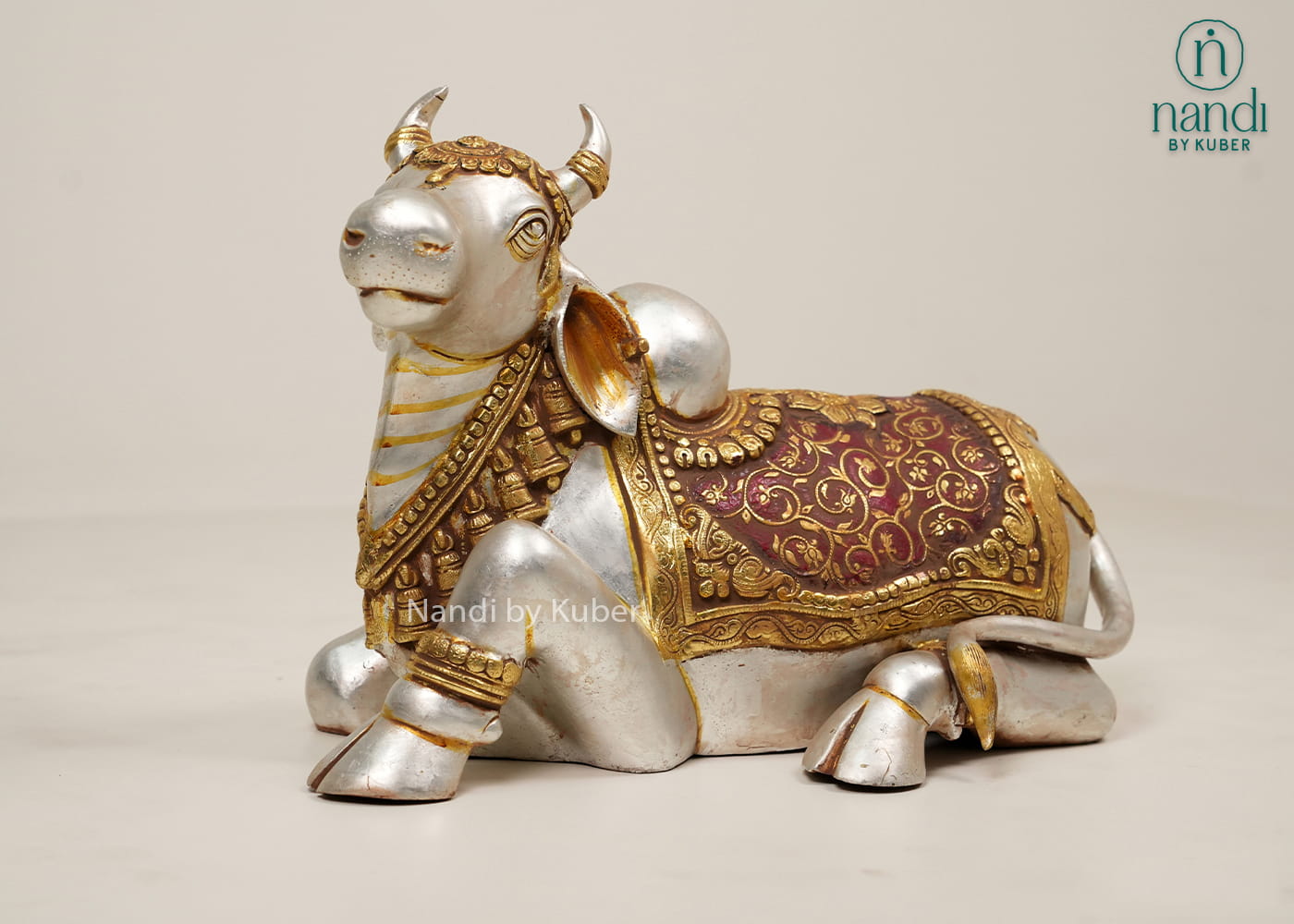 Silver and gold-tone Nandi bull statue with ornate carvings, decorative saddle cloth, and traditional seated design.