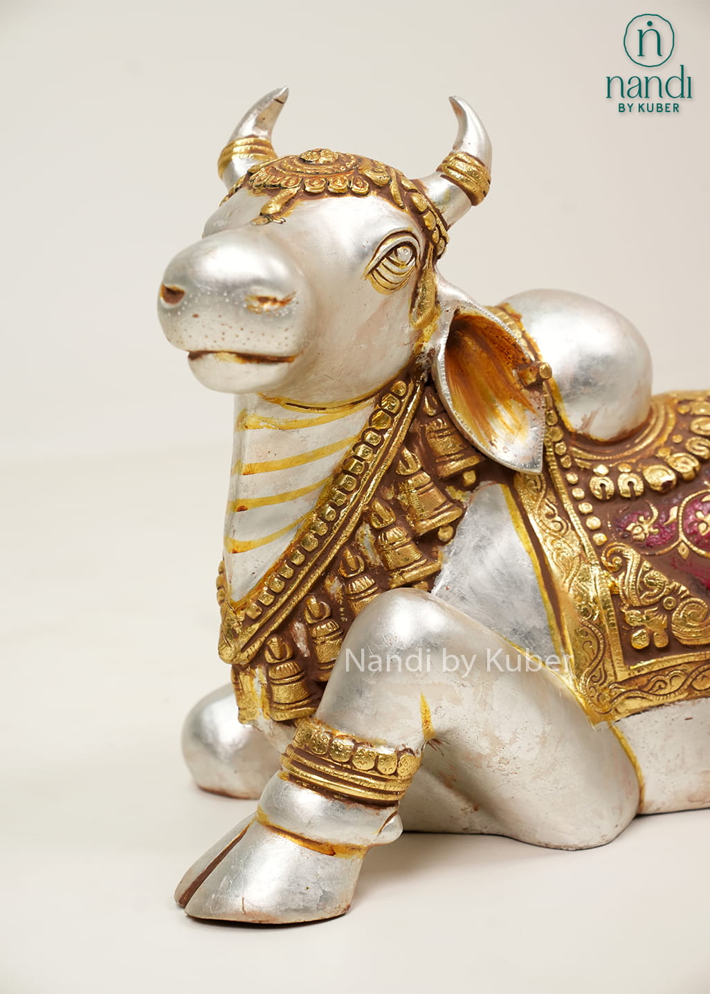 Silver and gold-tone Nandi bull statue with ornate carvings, decorative saddle cloth, and traditional seated design.